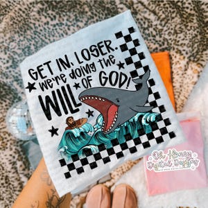 以下が含まれることがあります： 「GET IN, LOSER. we're doing the WILL OF GOD!」のテキストと、サメ、波、人物のグラフィックが描かれた水色のTシャツ。デザインには、白と黒のチェック柄と星が含まれています。