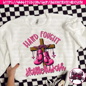 May include: White sweatshirt featuring a pink graphic design of a cross with boxing gloves hanging from it. The text "Hard Fought Hallelujah" is written in pink on the sweatshirt.