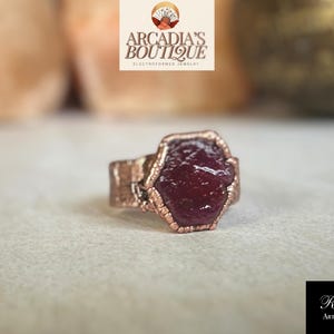 Pode incluir: Um anel de cobre com uma grande gema de forma irregular de cor vermelho escuro engastada no centro. O anel é uma joia eletroformada.