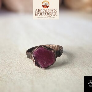 May include: A copper ring featuring a large, faceted, dark pink gemstone set within the band. The ring is handcrafted and has a rustic, organic appearance.