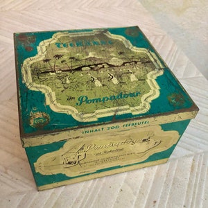 Può includere: Scatola di tè rettangolare vintage con una combinazione di colori verde acqua e oro. La scatola presenta un'illustrazione di paesaggio con palme e figure, e il testo "Tegernsee im Pompadour". La scatola è leggermente usurata, mostrando segni di età.