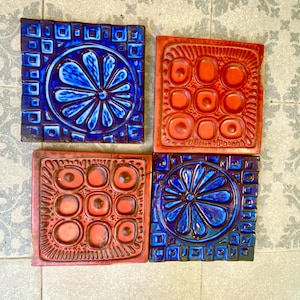 May include: Four square ceramic tiles arranged in a grid. Two tiles feature a deep blue glaze with a central floral motif surrounded by a geometric square pattern. Two tiles feature a vibrant orange glaze with a grid of nine circular indentations.