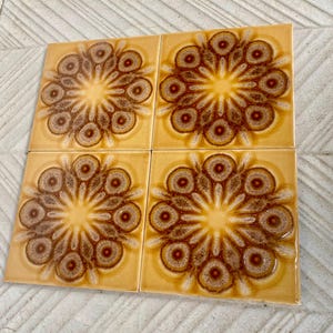 May include: Four square ceramic tiles with a repeating floral pattern. The tiles have a yellow background with a brown and tan floral design. The floral design has a central circle with radiating petals.