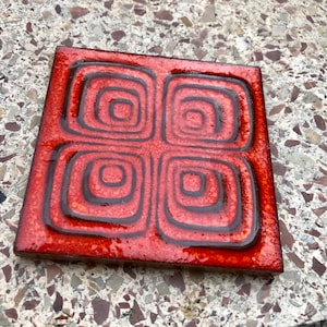 May include: A square ceramic tile with a glossy orange and red glaze featuring four embossed concentric square patterns in dark brown. The surface has a textured, mottled appearance with a reflective finish and raised edges defining the shapes.