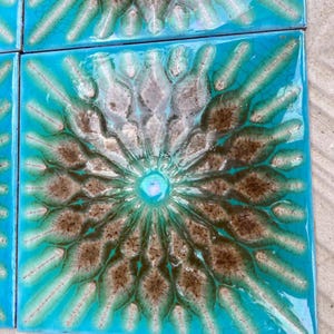 Op de afbeelding: Een vierkante keramische tegel met een glanzend turquoise glazuur en een centraal radiaal patroon. Het ontwerp bestaat uit verhoogde, getextureerde bruine en witte bloembladachtige vormen die uitstralen vanuit een klein, glad, lichtblauw middelpunt.