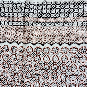 Op de afbeelding: Een witte stof met een herhalend patroon van bruine en zwarte geometrische vormen. De stof heeft een gegolfde rand.