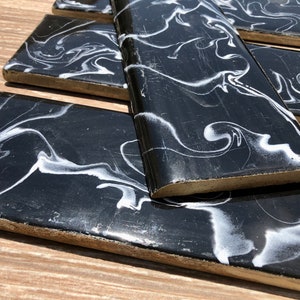 Op de afbeelding: Een verzameling rechthoekige zwarte tegels met een wit, wervelend marmerpatroon. De tegels hebben een glanzende afwerking en zijn gerangschikt op een houten oppervlak. De randen van de tegels zijn lichtbruin.