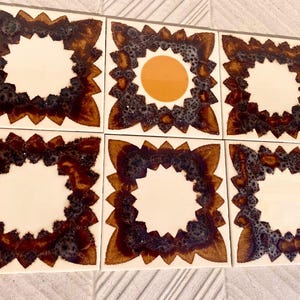 May include: Six square ceramic tiles with a repeating design. Each tile has a white centre surrounded by a brown, dark brown, and black floral-like pattern. One tile has an orange circle in the centre.