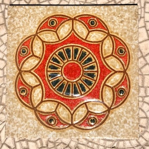 May include: Square ceramic tile featuring a symmetrical floral pattern. The centre is red with a black and blue sunburst design. The outer design is red with brown outlines and small green accents, set against a beige background.