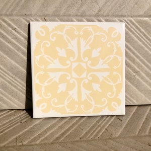 May include: A yellow ceramic tile with a white floral design. The tile is square shaped and has a smooth surface.