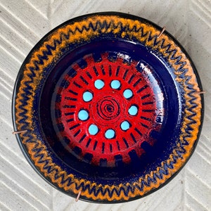 May include: A blue ceramic plate with a red and white sunburst design. The plate has a gold and orange rim with a zig-zag pattern.