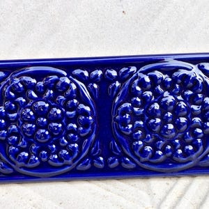 Könnte beinhalten: Rechteckige, kobaltblaue Keramikfliese mit erhabenem Blumendesign. Die Fliese zeigt zwei große, kreisförmige Blumenmuster mit kleineren, blasenartigen Details. Die glänzende Oberfläche verstärkt die komplizierten Details des geprägten Designs.