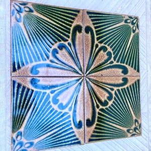 Peut inclure: Quatre carreaux de céramique carrés formant un grand carré. Chaque carreau présente un motif symétrique avec des teintes bleues, vertes et beiges. Le motif comprend des éléments floraux et en forme d'éventail stylisés, créant une surface décorative.