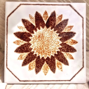 Può includere: Piastrella quadrata in ceramica con sfondo bianco e un motivo a girasole marrone. Il girasole ha petali marroni e un centro arancione maculato. La piastrella è incorniciata da un bordo marrone.