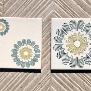 May include: Two white ceramic tiles featuring a geometric floral design. The design incorporates a stylised flower with a yellow centre and blue petals. The tiles are ideal for adding a touch of modern style to any space.