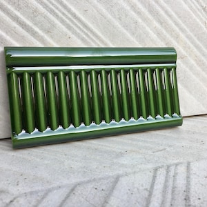 Könnte beinhalten: Eine rechteckige, glänzende, grüne Keramikfliese mit geripptem Design. Die Fliese hat eine abgerundete Oberkante und ist auf einer strukturierten, hellfarbigen Oberfläche angeordnet. Die Farbe der Fliese ist ein tiefes, sattes Grün.