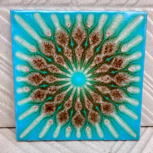 Op de afbeelding: Een vierkante keramische tegel met een levendige turquoise blauwe achtergrond en een centraal radiaal patroon. Het ontwerp bestaat uit bruin en wit gespikkelde bloembladachtige vormen die vanuit een turquoise middelpunt naar buiten stralen.