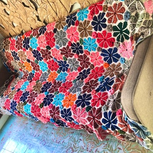 Peut inclure: Une couverture au crochet faite à la main, aux couleurs vives, ornée d'un motif floral, drapée sur un canapé. La couverture présente des couleurs variées : rouge, rose, bleu, orange et marron. Le canapé est gris neutre.