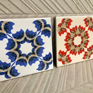 Può includere: Due piastrelle quadrate in ceramica con motivi floreali. Una piastrella presenta un motivo blu e beige su sfondo bianco. L'altra piastrella ha un motivo rosso e beige su sfondo bianco. Le piastrelle sono su una superficie strutturata.