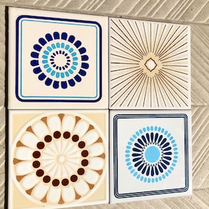 May include: Four square ceramic tiles with different geometric patterns. The tiles are white with blue, brown, and gold accents. The patterns include a circle with concentric rings, a sunburst design, a flower with petals and dots, and a circle with a blue center and concentric rings.