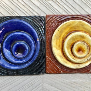 Peut inclure: Deux carreaux de céramique carrés avec un motif de cercles concentriques. Le carreau de gauche a un cercle bleu sur fond noir. Le carreau de droite a un cercle jaune sur fond marron.