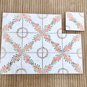 Può includere: Un insieme di piastrelle quadrate bianche con un motivo floreale. Ogni piastrella presenta fiori rosa e foglie verdi disposte in diagonale. Le piastrelle sono disposte su una superficie di legno.
