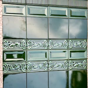Op de afbeelding: Een sectie groene keramische tegels met een glanzende afwerking. De tegels zijn gerangschikt in een rasterpatroon, met rechthoekige en vierkante vormen. Decoratieve bloemen- en cirkelpatronen zijn aanwezig in het ontwerp. De bovenrand heeft een geribbelde rand.