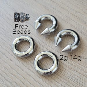 May include: Two silver-toned circular segment rings and two silver-toned horseshoe-shaped rings with pointed ends. The text Free Beads is near three small decorative beads, and the text 2g-14g is near the horseshoe rings.
