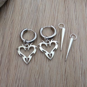 May include: Silver-toned hoop earrings with heart-shaped pendants and two spike charms. The heart pendants have a tribal design. The earrings and charms are displayed on a light brown wooden surface.
