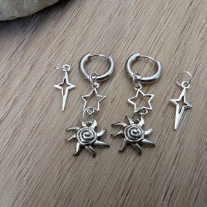 May include: Silver-toned earrings featuring celestial designs. The earrings include two pairs of hoop earrings with star and sun charms, and two single star-shaped studs. The sun charms have a spiral center and spiky rays.