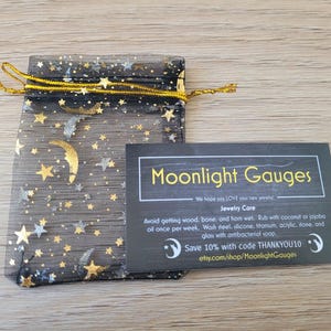 May include: A black mesh bag with gold stars and moons printed on it. The bag is tied with a gold ribbon. A black card with white text reads "Moonlight Gauges" and includes jewellery care instructions and a discount code.