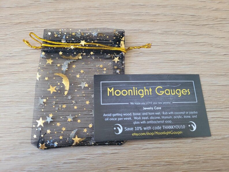 May include: A black mesh bag with gold stars and moons printed on it. The bag is tied with a gold ribbon. A black card with white text reads "Moonlight Gauges" and includes jewellery care instructions and a discount code.