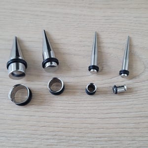 May include: A set of six silver ear gauges, including three pairs of plugs and three pairs of tapers. The tapers are pointed and the plugs are circular.