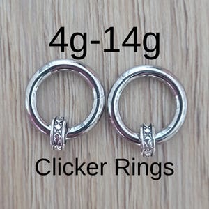 May include: Two silver clicker rings with decorative center pieces. The rings are labeled with the text "4g-14g" and "Clicker Rings". The jewelry is displayed on a light brown wooden surface.