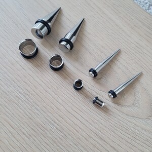 May include: A set of eight silver ear gauges and plugs, including a variety of sizes and styles. The gauges are made of metal and have a smooth, polished finish. The plugs are made of black rubber and have a flared design.