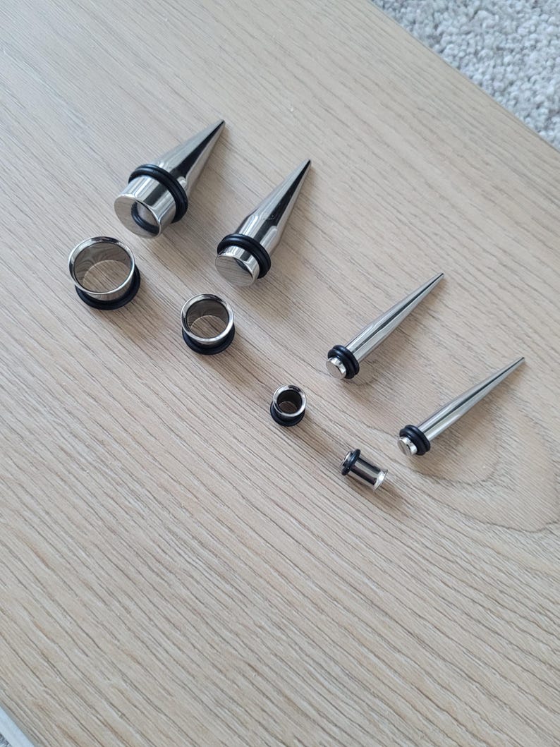 May include: A set of eight silver ear gauges and plugs, including a variety of sizes and styles. The gauges are made of metal and have a smooth, polished finish. The plugs are made of black rubber and have a flared design.
