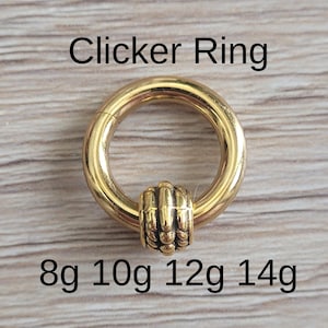 May include: A gold-colored clicker ring with a decorative center bead. The ring is circular with a hinged opening. Text on the image reads "Clicker Ring" and "8g 10g 12g 14g".
