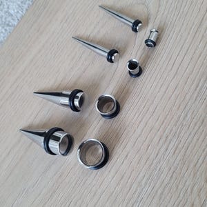 May include: A set of six silver ear gauges, including three pairs of plugs and three pairs of tapers. The tapers are pointed on one end and have a flared end on the other. The plugs are circular and have a black rubber insert.