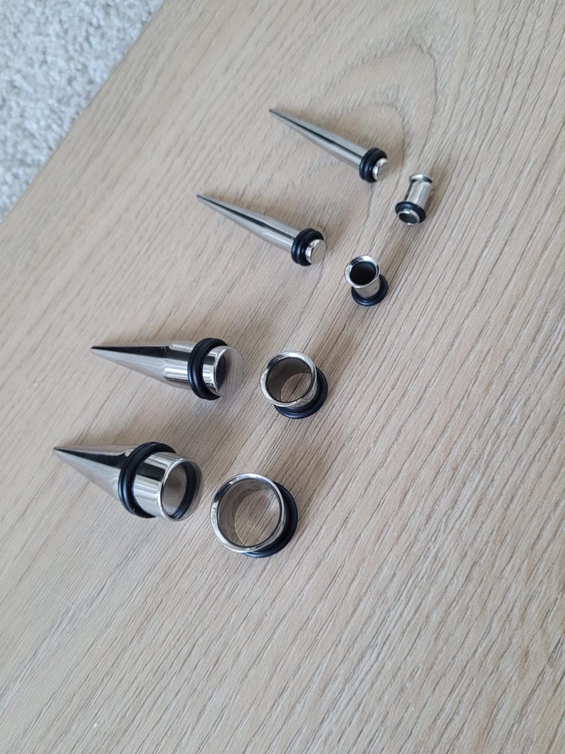 May include: A set of six silver ear gauges, including three pairs of plugs and three pairs of tapers. The tapers are pointed on one end and have a flared end on the other. The plugs are circular and have a black rubber insert.