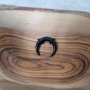 May include: A black crescent moon shaped ear gauge, made of acrylic, is displayed on a wooden surface.