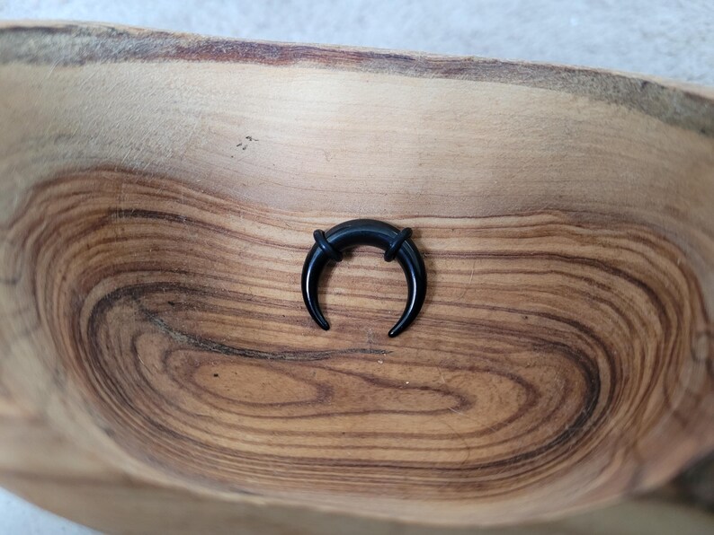 May include: A black crescent moon shaped ear gauge, made of acrylic, is displayed on a wooden surface.
