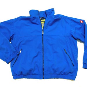 Puede incluir: Chaqueta azul brillante con cremallera completa, dos bolsillos laterales y un pequeño logotipo en la manga superior. La chaqueta tiene un cuello alto y puños elásticos. La chaqueta está hecha de un material ligero y resistente al agua.