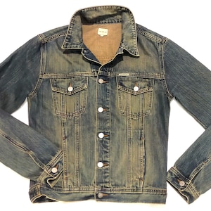 Può includere: Giacca di jeans blu con un design classico. Presenta una chiusura con bottoni, due tasche sul petto e due tasche laterali. Il denim ha un aspetto sbiadito e usurato. Il colletto e la fodera interna sono di colore marrone chiaro.