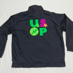 May include: Black nylon jacket with long sleeves. The back features the text "US OPEN" in green, pink, and orange, with a tennis ball graphic. The jacket has a high collar and a full zipper.