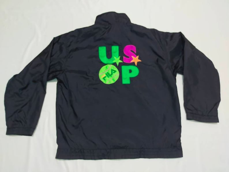 May include: Black nylon jacket with long sleeves. The back features the text "US OPEN" in green, pink, and orange, with a tennis ball graphic. The jacket has a high collar and a full zipper.