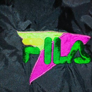 May include: Close-up of a black athletic jacket with a colorful embroidered logo. The logo features the word "FILA" in green, with a purple and lime green design. The jacket appears to be made of a shiny, smooth material.