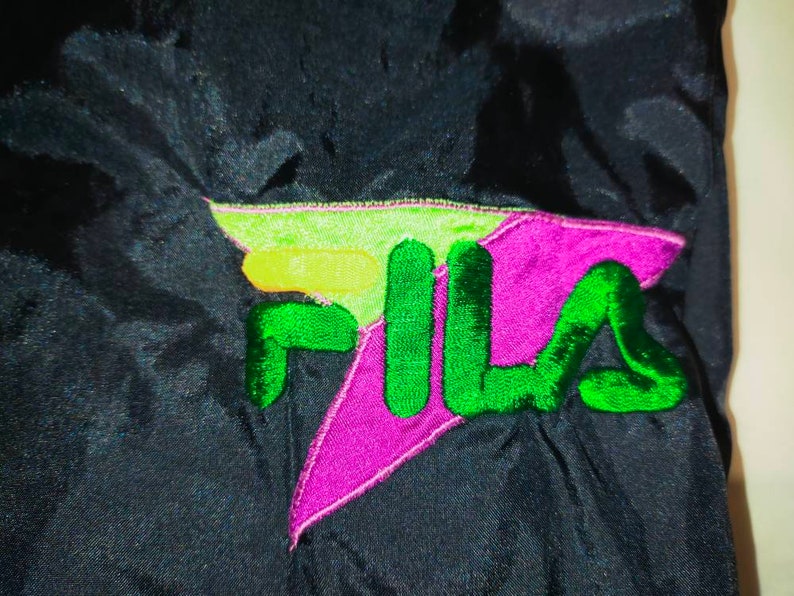 May include: Close-up of a black athletic jacket with a colorful embroidered logo. The logo features the word "FILA" in green, with a purple and lime green design. The jacket appears to be made of a shiny, smooth material.