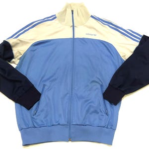Può includere: Giacca da tuta con zip blu chiaro e blu navy con dettagli bianchi. La giacca ha un collo alto, due tasche anteriori e il logo Adidas. Le maniche sono blu navy e le spalle hanno strisce bianche.