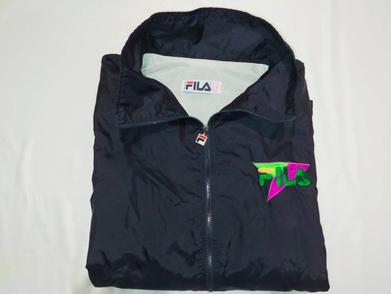 May include: A navy blue FILA windbreaker jacket with a full zipper and a high collar. The jacket features the FILA logo on the chest in green, pink, and yellow. The FILA logo is also on the tag inside the jacket.