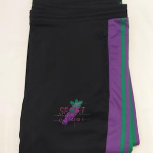 Può includere: Pantaloni sportivi neri con una striscia viola e verde sul lato. I pantaloni hanno un girovita con coulisse e un piccolo logo con la scritta "SPORT" e il logo Adidas in viola e verde.